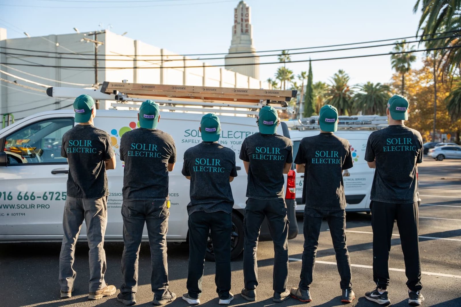 Solir Electric crew seen from behind in branded work shirts beside service vehicles